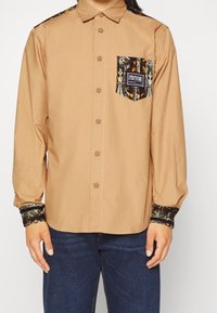 Brun långärmad skjorta med en mönstrad ficka, som har mönstrade ärmslut och axlar. Knappstängning och mjukt, bomullsmaterial.