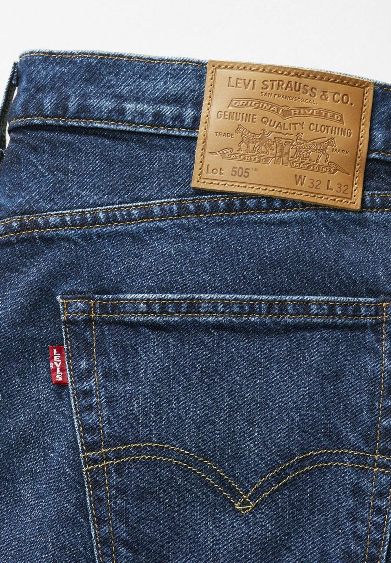 Jeans aus Denim von Levi Strauss & Co., mit einem braunen Lederpatch mit eingeprägtem Logo, blauer Stoff, goldfarbene Nähte und rotes Tab-Detail.
