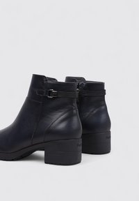 Botas de tobillo de cuero negro con punta redonda, cierre de cremallera lateral, correa decorativa con hebilla y un tacón bajo y grueso. Textura suave.