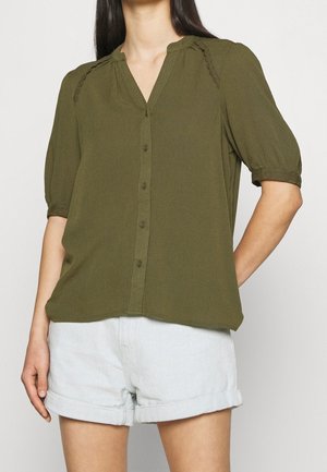Blusa a maniche corte verde oliva con colletto a punta, chiusura con bottoni frontale e dettagli testurizzati sulle spalle; abbinata a pantaloni corti in denim chiaro.
