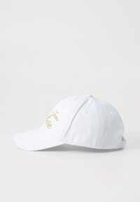 White baseball cap made of cotton fabric with a curved brim. Features gold embroidered text on the front and adjustable strap at the back.