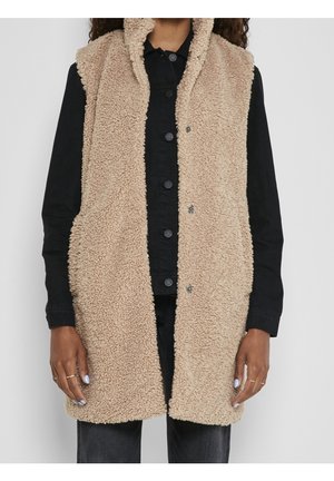 Femme portant un gilet en polaire beige sans manches boutonné, sur un t-shirt noir à manches longues et un pantalon foncé, debout les bras relâchés.