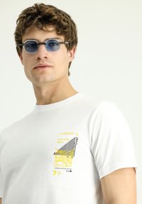 Camiseta blanca de algodón con un diseño gráfico colorido que incluye elementos amarillos y negros. Acompañada de unas gafas de sol redondas azules con detalles negros.
