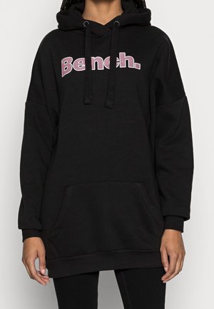 Personne portant un sweat à capuche noir oversized avec « Bench. » en lettres violettes et blanches sur la poitrine, poche avant et cordons pendants.