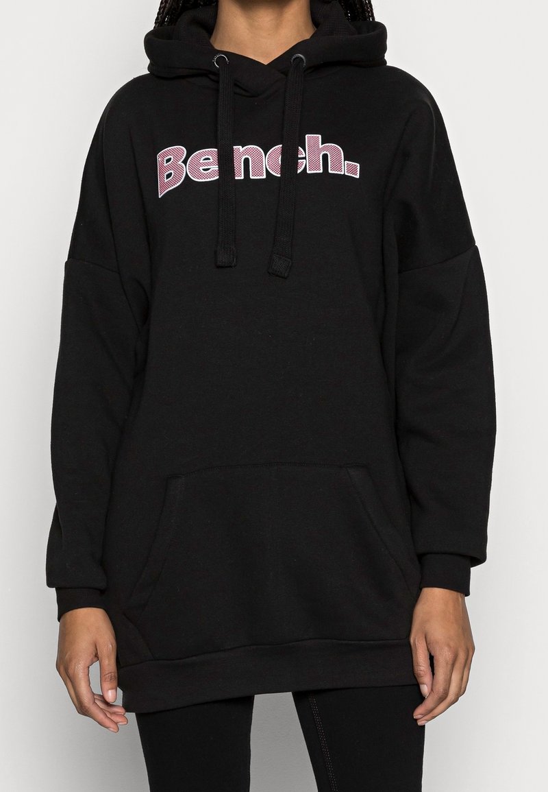 Personne portant un sweat à capuche noir oversized avec « Bench. » en lettres violettes et blanches sur la poitrine, poche avant et cordons pendants.
