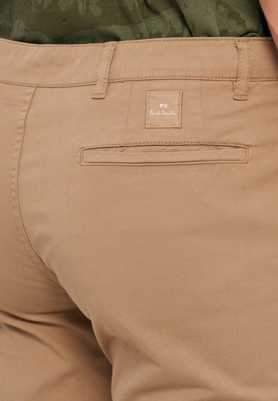 Pantaloni in cotone color sabbia con una tasca posteriore caratterizzata da un'etichetta cucita con la scritta "PS Paul Smith". Texture liscia, taglio dritto e design minimal.