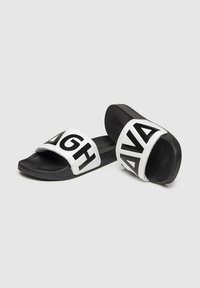 Chanclas en blanco y negro con una correa ancha que presenta grandes letras negras sobre un fondo blanco. Suela plana con superficie texturizada.