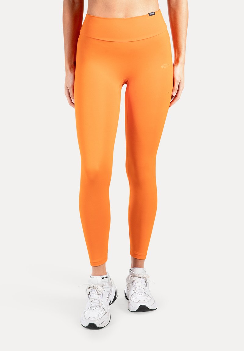 Orangea leggings med hög midja, slät och stretchig textur, figurnära design och en diskret logodetalj nära midjebandet, matchade med vita sneakers.