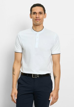 Man met kort krullend haar, gekleed in een wit poloshirt met korte mouwen en een marineblauwe broek, staand tegen een effen lichtgrijze achtergrond.