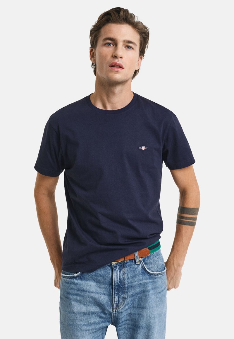 Camiseta de algodón azul marino con cuello redondo, que presenta un pequeño logo en el lado izquierdo del pecho y mangas cortas. Lleva puesta unos jeans de color azul claro.