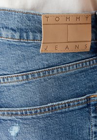 Jeans de mezclilla en azul claro con un leve desgaste. Presentan una etiqueta de cuero marrón con "TOMMY JEANS" embellecida, mostrando detalles de costura.