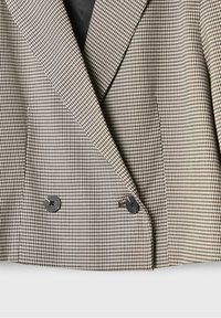 Double-breasted blazer in a houndstooth pattern, featuring brown and cream colors, sharp lapels, and black buttons. Textured fabric.
