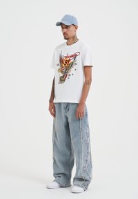 Bílý bavlněný tričko s barevným grafikou orla a textem "97"; kombinováno se světle modrými oversized džíny s boční výšivkou a potrhanými okraji.