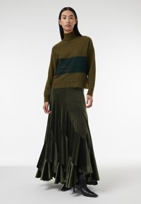 Groene gebreide trui met een donkergroene horizontale streep, gecombineerd met een gelaagde olijfgroene fluwelen maxi-rok en zwarte hakboots.