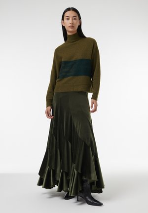 Groene gebreide trui met een donkergroene horizontale streep, gecombineerd met een gelaagde olijfgroene fluwelen maxi-rok en zwarte hakboots.
