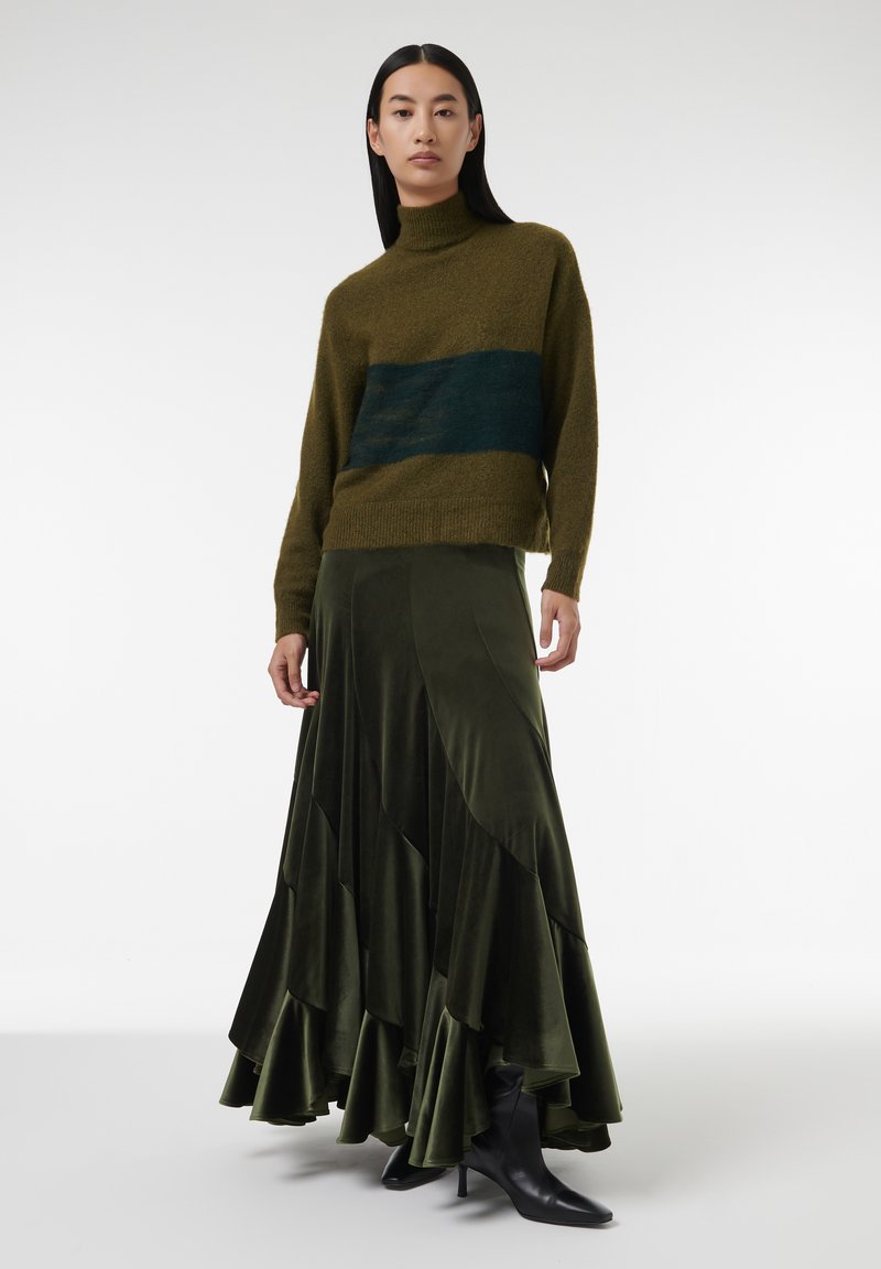 Groene gebreide trui met een donkergroene horizontale streep, gecombineerd met een gelaagde olijfgroene fluwelen maxi-rok en zwarte hakboots.