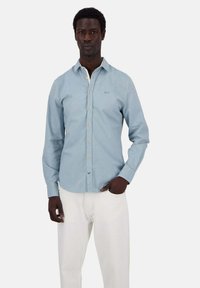 Camicia azzurra chiara con colletto, maniche lunghe e un logo ricamato discreto sul petto; abbinata a pantaloni bianchi.