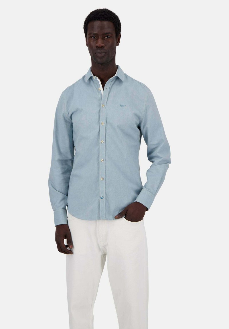 Camicia azzurra chiara con colletto, maniche lunghe e un logo ricamato discreto sul petto; abbinata a pantaloni bianchi.