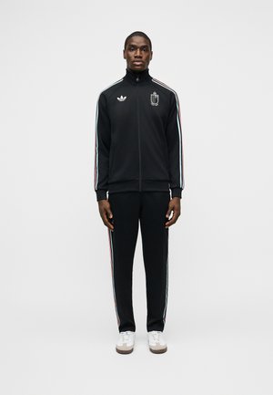 Hombre de pie, vestido con chaqueta y pantalones deportivos negros de Adidas con franjas rojas, blancas y azules en las mangas y las piernas, y zapatillas blancas.