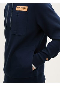 Sweatshirt marine avec une poche poitrine zippée, des poignets côtelés et une grande poche latérale. Équipé de cordons en noir et blanc et d'un logo orange.