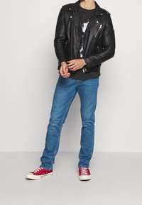 Giacca da motociclista in pelle nera, jeans in denim blu e sneakers rosse alte. La giacca presenta cerniere e un design aderente.