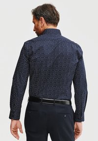 Granatowa koszula zapinana na guziki w białe kropki, z długimi rękawami i kołnierzykiem rozłożystym. Noszona z czarnymi spodniami i czarnym paskiem.
