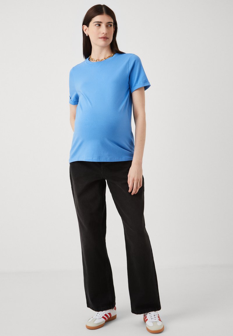 Blauwe katoenen t-shirt met korte mouwen, gecombineerd met zwarte wijde broek. Model draagt witte sneakers met rode accenten. Eenvoudige, casual stijl.