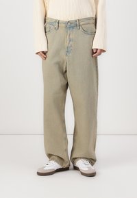 Jeans beige de pierna ancha con un acabado desgastado, que cuentan con cierre de botón y cremallera, bolsillos frontales y un ajuste relajado, combinados con zapatillas blancas.