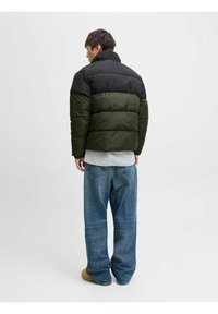 Giacca imbottita con la parte superiore nera e la parte inferiore verde oliva; presenta un colletto alto e una vestibilità oversize. Indossata sopra una camicia a quadri e jeans di denim.