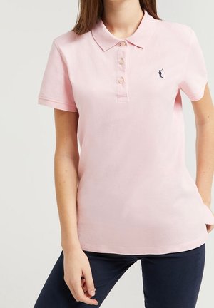Kobieta nosząca jasnoróżową koszulkę polo z krótkim rękawem, trzema guzikami i małym, ciemnym, haftowanym logo na lewej piersi.