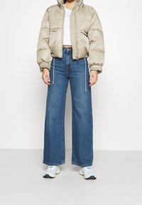 Beige padded jacket with a hood, elastic cuffs, and two front pockets, paired with high-waisted wide-leg blue jeans and white sneakers.