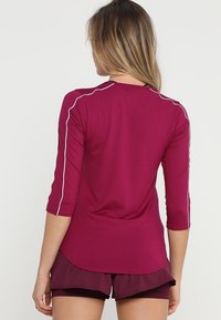 Vinröd träningströja med trekvartslånga ärmar och vita kontrasterande sömmar, tillverkad av ett slätt, lätt material.