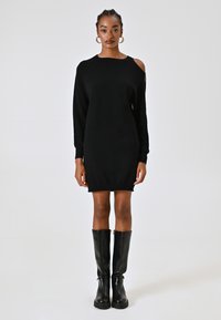 Donna con capelli intrecciati indossa un vestito di maglia nero a maniche lunghe con spacco sulle spalle e stivali neri al ginocchio.