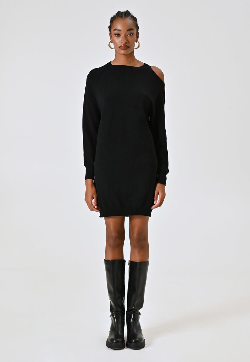 Donna con capelli intrecciati indossa un vestito di maglia nero a maniche lunghe con spacco sulle spalle e stivali neri al ginocchio.