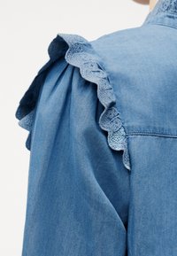 Μπλε τζιν ένδυμα με βολάν δαντέλα στο μανίκι και λεπτομέρεια με μαζεμένο ύφασμα κάτω από το βολάν.