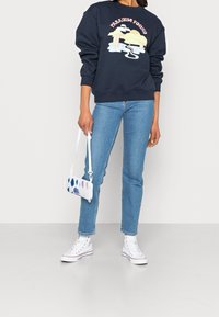 Námořnická mikina s grafikou "Paradise Found", světle modré džíny a bílé tenisky. Doplňěk: malá vícerozměrná taška na popruhu.