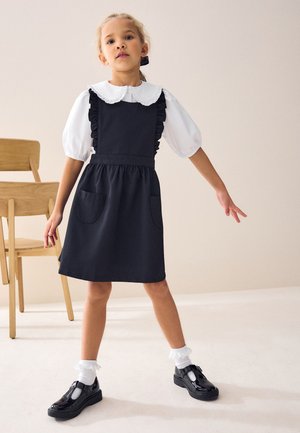 Jeune fille debout en robe tablier noire avec poches, chemisier blanc à manches bouffantes, chaussettes à volants et chaussures noires, à côté de deux chaises en bois.