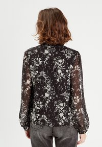 Blouse noire à motifs floraux avec un tissu transparent, dotée de manches longues, d'une légère bouffante à l'épaule et d'un délicat imprimé de fleurs blanches.