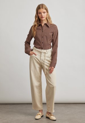 Junge Frau mit langen blonden Haaren, die ein braunes kariertes Hemd, cremefarbene weite Hosen und beige Loafer trägt, steht mit einer Hand in der Tasche.