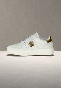 Λευκό sneaker χαμηλού ύψους με λευκά κορδόνια, χρυσή ταμπέλα στη φτέρνα και χρυσό διακοσμητικό λογότυπο στο πλάι, εμφανιζόμενο σε φωτεινή υφασμάτινη επιφάνεια.