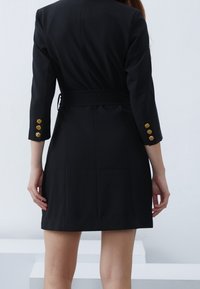 Kvinna iklädd en svart kavajklänning med bälte och tre guldknappar på varje ärm, visad från baksidan mot en neutral ljus bakgrund.