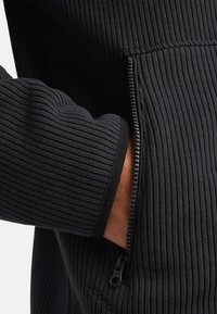 Jogger in tessuto nero a coste con tasca con zip. Caratterizzato da una superficie testurizzata e polsini elasticizzati, progettato per comfort e funzionalità.