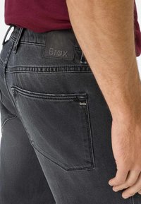 Jeans in denim grigio scuro con una finitura testurizzata, dotati di un'etichetta in pelle nera riportante la scritta "Brax" sulla vita e in una tasca posteriore.