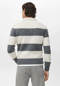 Hombre con cabello rizado que lleva un suéter de rayas anchas en gris y blanco y jeans grises, de pie de espaldas frente a un fondo blanco.