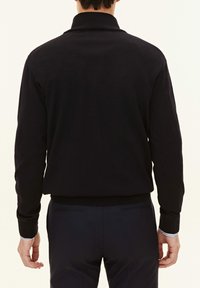 Svart stickad pullover med hög krage, ribbvävd struktur och långa ärmar. Avslutad med ett tajt midjeband, i kombination med mörka byxor.
