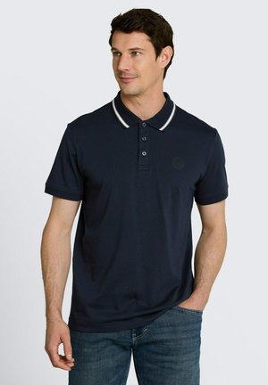 Mężczyzna ubrany w granatową koszulkę polo z krótkim rękawem z białym obszyciem kołnierzyka, niebieskie jeansy, patrzy lekko w lewo z jedną ręką w kieszeni.