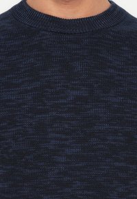 Close-up of a person wearing a dark blue and black textured knit sweater with a ribbed crew neck collar.