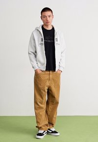 Grijze hoodie met een rits aan de voorkant, zwart graphic t-shirt, bruine corduroy broek en zwarte sneakers met witte accenten.