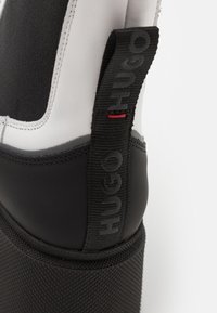 Botte en caoutchouc noire et blanche avec semelle texturée et languette en tissu noir avec marquage en relief "HUGO" et une petite couture rouge.