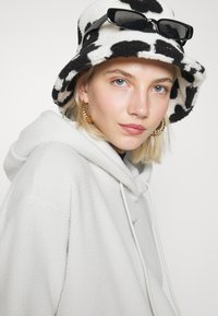 Fleecehuvtröja i ljusgrått med dragsnörning. Bär en svart och vit cow-print bucket hat tillsammans med solglasögon och guld hoopsörhängen.